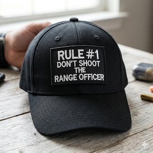 May include: Black baseball cap with a curved brim. The cap features a rectangular patch with the text "RULE #1 DON'T SHOOT THE RANGE OFFICER" in white lettering. The cap is made of a durable fabric and has a structured fit.