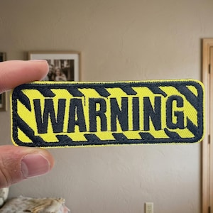May include: A rectangular patch with the word "WARNING" in bold black letters on a bright yellow background. The patch has a black border with a diagonal stripe design. The patch is held by a person's fingers.