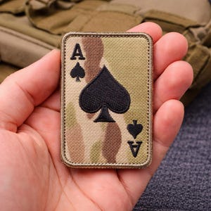 May include: A rectangular patch featuring the Ace of Spades playing card design. The patch has a camouflage background with the black spade symbol and the letter "A" in black. The edges are stitched with a tan thread.