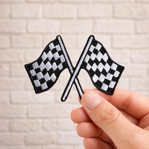 May include: Two embroidered checkered racing flags, black and white, are crossed. The flags are held against a light-colored brick background. The flags are a classic symbol of racing and speed.