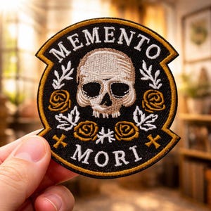 May include: Embroidered patch with a skull design, surrounded by roses and leaves. The patch features the words "MEMENTO MORI" in white lettering on a black background, with gold and white accents.