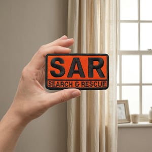 May include: An orange rectangular patch with the letters "SAR" in bold black lettering. Below the letters, the words "SEARCH & RESCUE" are embroidered in black. The patch is held in front of a window with a beige curtain.