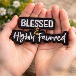 May include: Two black embroidered patches with white text. The top patch reads "BLESSED" and the bottom patch reads "Highly Favored" with a gold ampersand. The patches are held in open hands.