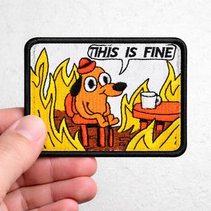 May include: Embroidered patch featuring a cartoon dog sitting in a burning room, with the text "THIS IS FINE" in a speech bubble. The dog is orange with a red hat, and the flames are yellow and orange. The patch has a black border.