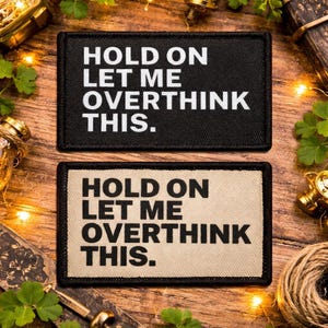 May include: Two rectangular patches with the text "HOLD ON LET ME OVERTHINK THIS." The top patch is black with white text, and the bottom patch is tan with white text. The patches are on a wooden surface with decorative items.