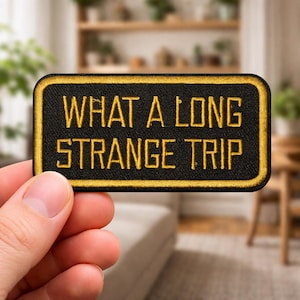 May include: Black rectangular patch with gold embroidered border and text. The text reads "WHAT A LONG STRANGE TRIP" in gold block letters. The patch is held in a person's hand.