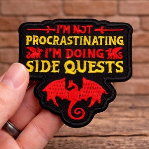 May include: Black embroidered patch with the text "I'M NOT PROCRASTINATING I'M DOING SIDE QUESTS" in yellow. The patch features red dragon illustrations and accents.