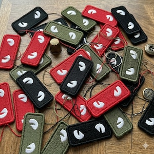 May include: A collection of embroidered patches in red, black, and olive green. Each patch is rectangular with a white, curved design. A thimble, thread, and buttons are also visible.