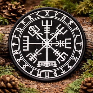 May include: A round, black patch with a white, intricate Viking symbol design. The patch features a circular border with runes and a central compass-like emblem. The patch is surrounded by pine needles and pine cones.