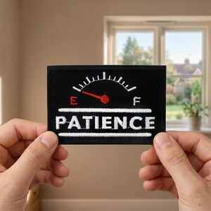 May include: Black rectangular patch with the word "PATIENCE" embroidered in white. Above the word is a gauge with a red needle pointing towards "E". The patch is held up by two hands.