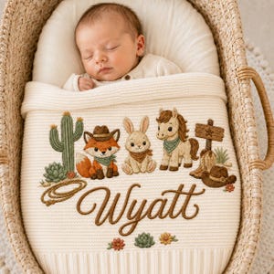 Könnte beinhalten: Eine cremefarbene Strickdecke für Babys mit gestickten Western-Motiven, darunter ein Fuchs mit Cowboyhut, ein Hase, ein Pferd, ein Kaktus und ein Paar Stiefel. Der Name Wyatt ist mittig in brauner Schreibschrift aufgestickt.