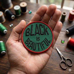 May include: A round, green patch with the words "BLACK IS BEAUTIFUL" embroidered in black. The patch has a red border and is held in a hand. Sewing supplies, including spools of thread and scissors, are in the background.