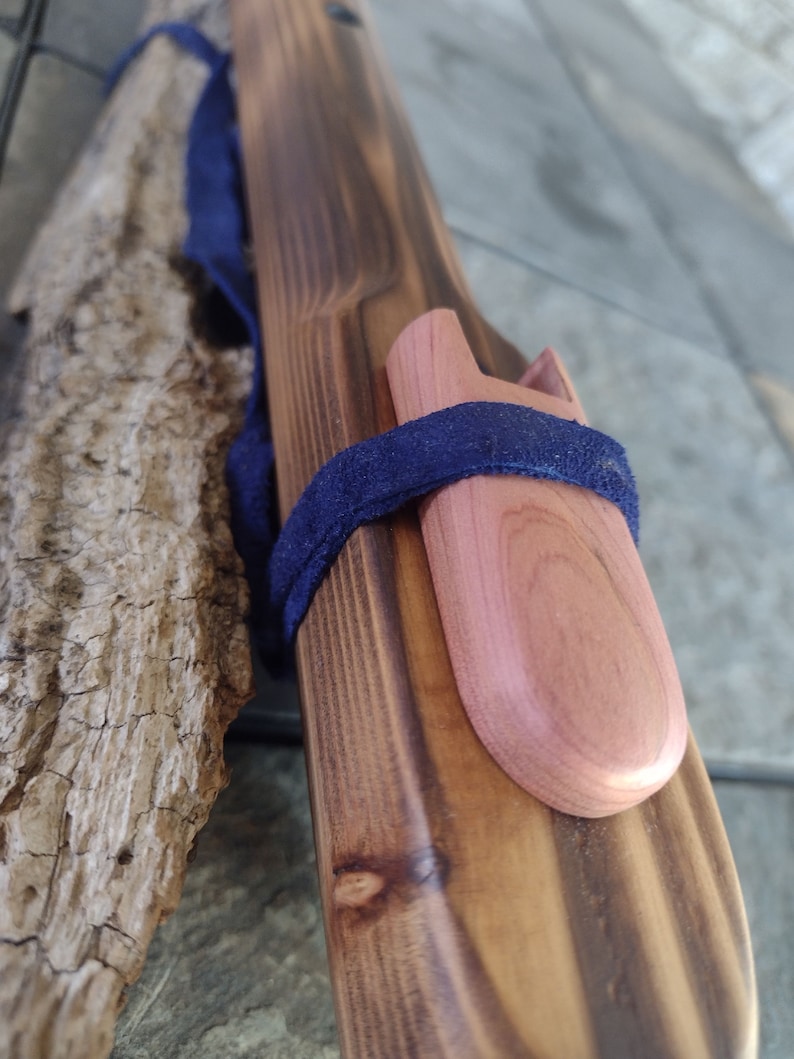 May include: A wooden flute with a dark brown finish and a natural wood mouthpiece. The flute is wrapped with a dark blue strap and has a small, round, light brown wooden plug on the end.