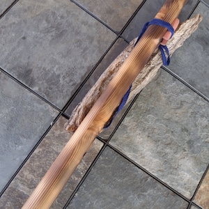 May include: A long, wooden flute with a dark brown stain. The flute is tied with a blue cord and rests on a piece of driftwood.