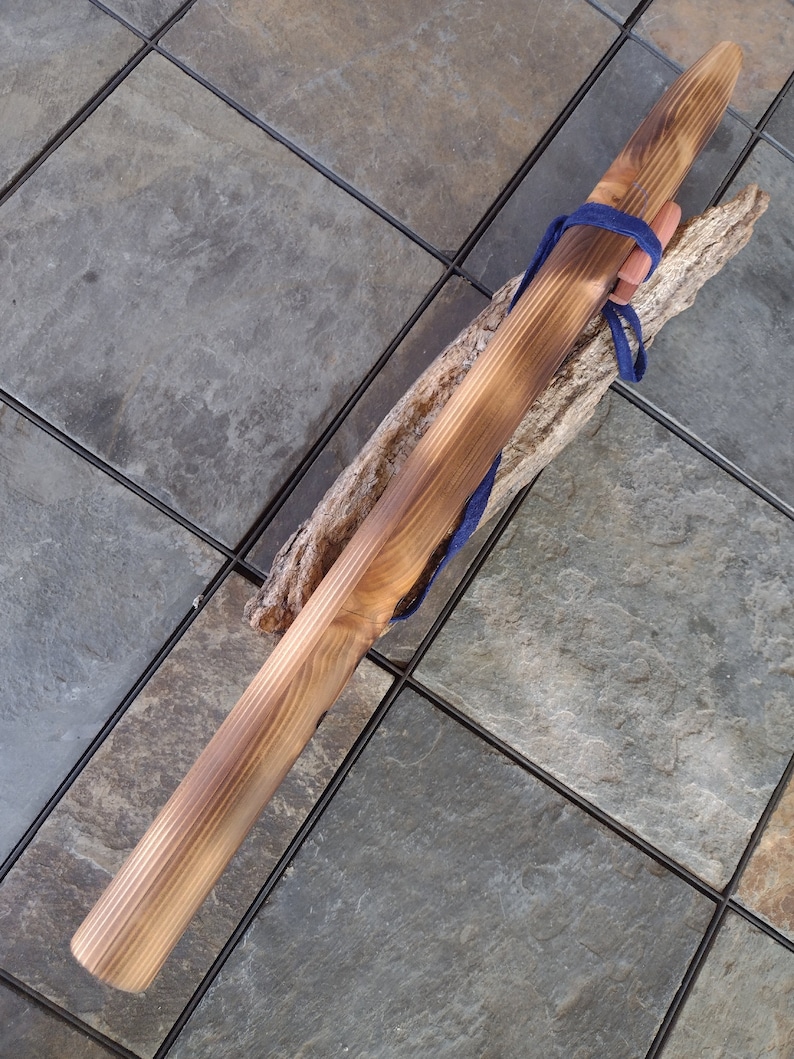 May include: A long, wooden flute with a dark brown stain. The flute is tied with a blue cord and rests on a piece of driftwood.