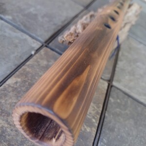 May include: A wooden flute with a dark brown stain. The flute has multiple finger holes and is resting on a piece of driftwood.