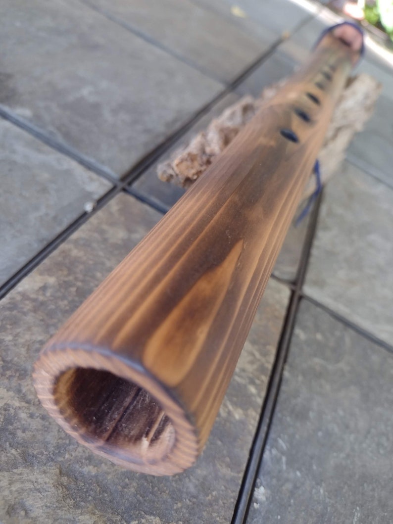 May include: A wooden flute with a dark brown stain. The flute has multiple finger holes and is resting on a piece of driftwood.