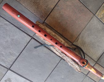 May include: A handcrafted wooden flute with a reddish-brown finish. It features decorative carvings, a turquoise accent, and black finger holes. A black leather strap is attached. The flute rests on a piece of wood.