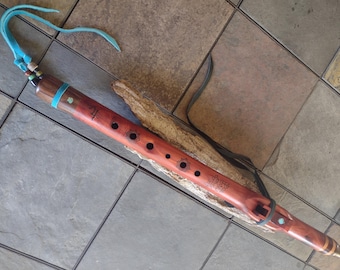 May include: A handcrafted wooden flute with a reddish-brown finish, featuring several finger holes and decorative turquoise accents. The flute has a blue leather strap and is resting on a piece of wood. The instrument is designed with a Native American aesthetic.
