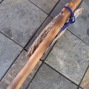 May include: A wooden flute with a dark brown stain and a blue cord tied around it. The flute is resting on a piece of driftwood.