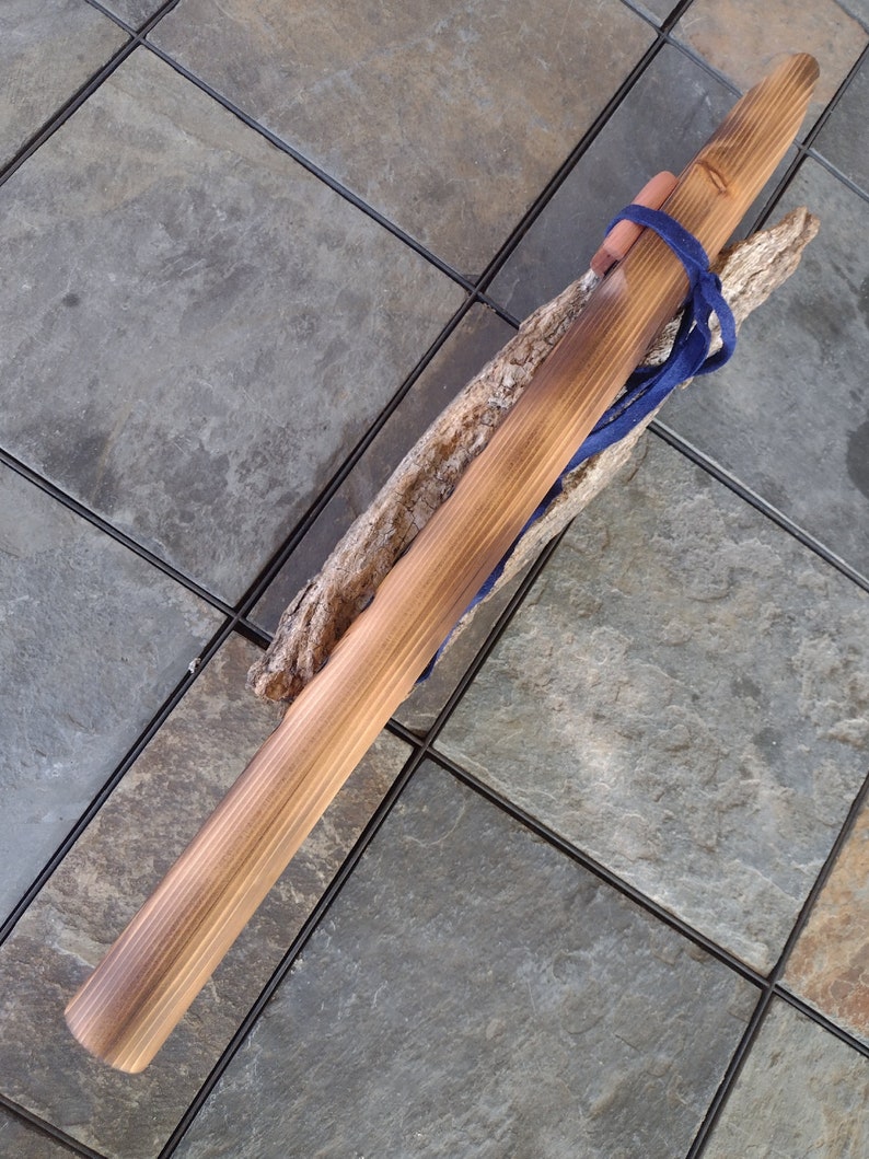 May include: A wooden flute with a dark brown stain and a blue cord tied around it. The flute is resting on a piece of driftwood.