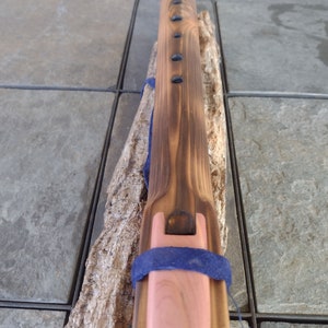 May include: A wooden flute with a dark brown stain and a pink mouthpiece. The flute has a blue strap and is decorated with black dots.