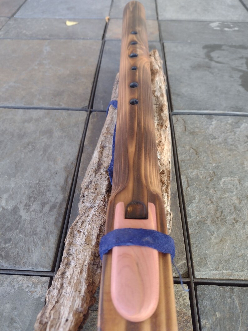 May include: A wooden flute with a dark brown stain and a pink mouthpiece. The flute has a blue strap and is decorated with black dots.