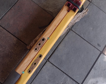 May include: A handcrafted wooden flute with a light yellow body and a reddish-brown mouthpiece. The flute features decorative accents, including turquoise-colored stones and a dark brown band. The instrument rests on a piece of wood against a tiled surface.