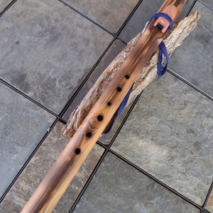 May include: A wooden Native American flute with a dark brown stain and a blue leather strap. The flute has six finger holes and a mouthpiece at one end. The flute is resting on a piece of driftwood.