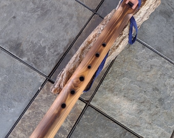 May include: A wooden Native American flute with a dark brown stain and a blue leather strap. The flute has six finger holes and a mouthpiece at one end. The flute is resting on a piece of driftwood.