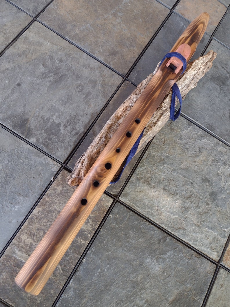 May include: A wooden Native American flute with a dark brown stain and a blue leather strap. The flute has six finger holes and a mouthpiece at one end. The flute is resting on a piece of driftwood.