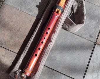 May include: A handcrafted wooden flute with a reddish-brown finish, featuring a light-colored wood accent near the mouthpiece. The flute has several finger holes and is encased in a soft, gray carrying case with a strap. The instrument is designed for musical performance.