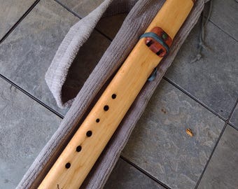 May include: A wooden flute with a light brown finish, featuring decorative carvings and a green stone inlay. The flute rests in a gray, soft case. The flute has a dark red end cap and a series of finger holes.