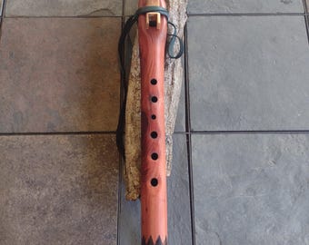 May include: A handcrafted wooden flute with a reddish-brown finish, featuring a light-colored mouthpiece and decorative accents. The flute has several finger holes and is adorned with a leather strap and a tan leather cord. The instrument rests on a piece of textured wood.