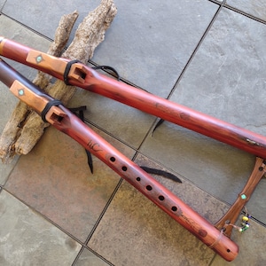 Peut inclure: Deux flûtes en bois faites à la main, dans des tons bruns et brun-rouge, avec des accents décoratifs. Une flûte porte les initiales "HC" gravées. Les flûtes reposent sur une branche en bois clair et une surface carrelée.