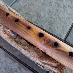 May include: A wooden flute with six finger holes, resting on a piece of driftwood. The flute is brown with a natural wood finish.