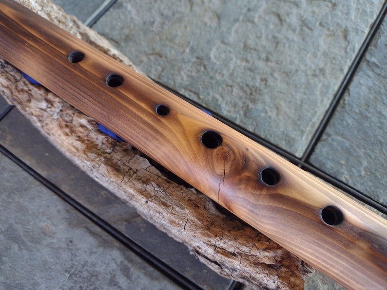 May include: A wooden flute with six finger holes, resting on a piece of driftwood. The flute is brown with a natural wood finish.