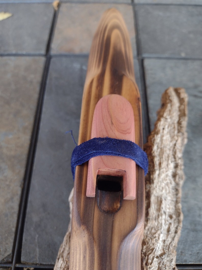 May include: A wooden flute with a dark brown stain and a natural wood mouthpiece. The flute is decorated with a blue ribbon and a red wooden cap.