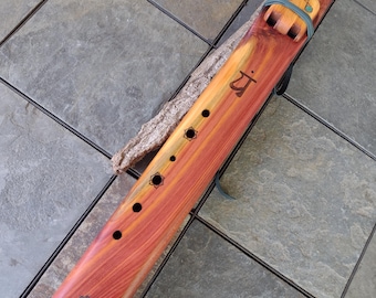 May include: A handcrafted wooden flute with a reddish-orange and brown finish. The flute features decorative turquoise accents, a leather strap, and a carved design near the mouthpiece. The instrument has several finger holes and a unique, textured end cap.