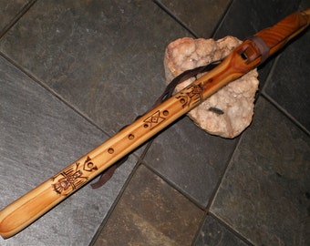 May include: A handcrafted wooden flute with intricate etched designs and a leather strap. The flute is light brown with darker brown etched patterns and is resting on a white and tan stone.