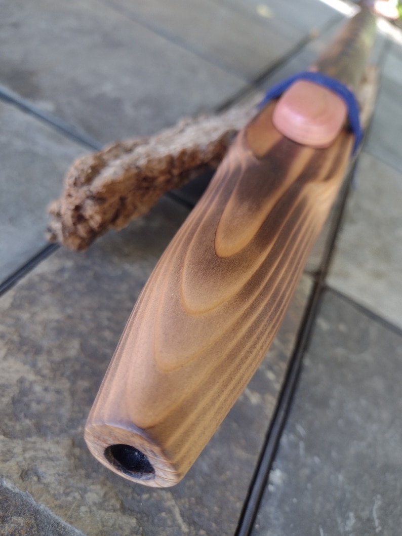 May include: A wooden flute with a brown stain and a natural wood finish. The flute has a small hole at the end and a blue string wrapped around the top.