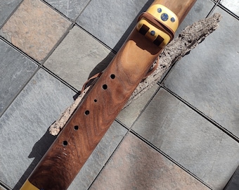 May include: A handcrafted wooden flute with a warm brown finish, featuring decorative accents and a leather strap. The flute has several finger holes and a light-colored mouthpiece. The instrument rests on a piece of bark.