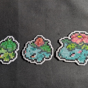 May include: Three cross-stitched Bulbasaur designs in various stages of evolution. The first is green, the second is blue with a red bulb, and the third has a pink and yellow flower. Each has a gray border and is set against a dark background.