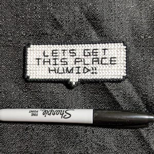 Könnte beinhalten: Ein weiß-schwarzer Kreuzstich-Patch mit dem Text "LETS GET THIS PLACE HUMID!!" liegt auf einer strukturierten grauen Oberfläche. Unter dem Patch befindet sich ein schwarz-weißer Sharpie-Feinmarker.