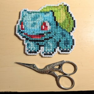May include: A cross-stitch of a Bulbasaur character in shades of blue, green, red, and white. The Bulbasaur is on a light brown surface next to a pair of silver embroidery scissors.