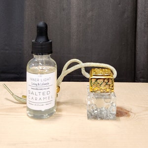 May include: A clear glass bottle with a gold cap and a gold string attached to a clear glass bottle with a gold top. The bottle is labeled "Inner Light Living & Lifestyle" and "Salted Caramel Reed Diffuser Refill".