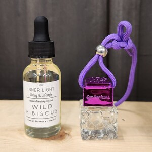 May include: A clear glass bottle with a black dropper top and a white label that reads "INNER LIGHT Living & Lifestyle treasuredbytrinity.etsy.com WILD HIBISCUS Reed Diffuser Refill". A purple car air freshener with a silver metal top and a clear glass base. The air freshener is attached to a purple string.
