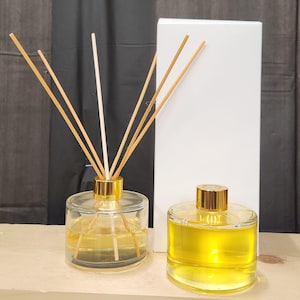 May include: Two glass aromatherapy diffusers with gold accents. One diffuser has wooden reeds inserted, the other has a gold cap. Both contain a yellow liquid. A white box is in the background.