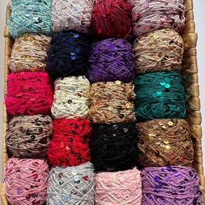 May include: A woven basket filled with various colorful yarn balls. The yarn is in shades of teal, pink, red, black, and brown, with sequins. The basket has a brown handle. The yarn is suitable for knitting and crochet projects.