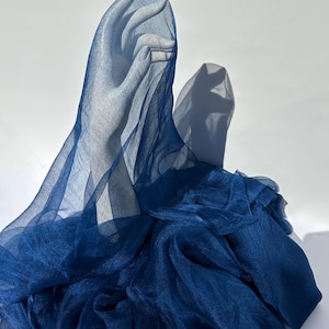 Peut inclure: Un tissu bleu marine profond et transparent drapé sur un présentoir en forme de main de couleur claire. La matière translucide crée des plis et des couches souples, le tissu masquant partiellement la forme tout en projetant une ombre.